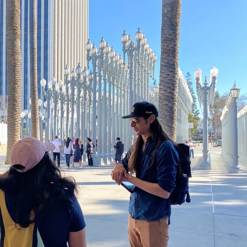 Two people stand near a large outdoor installation of numerous street lamps. A tall building and palm trees are in the background.