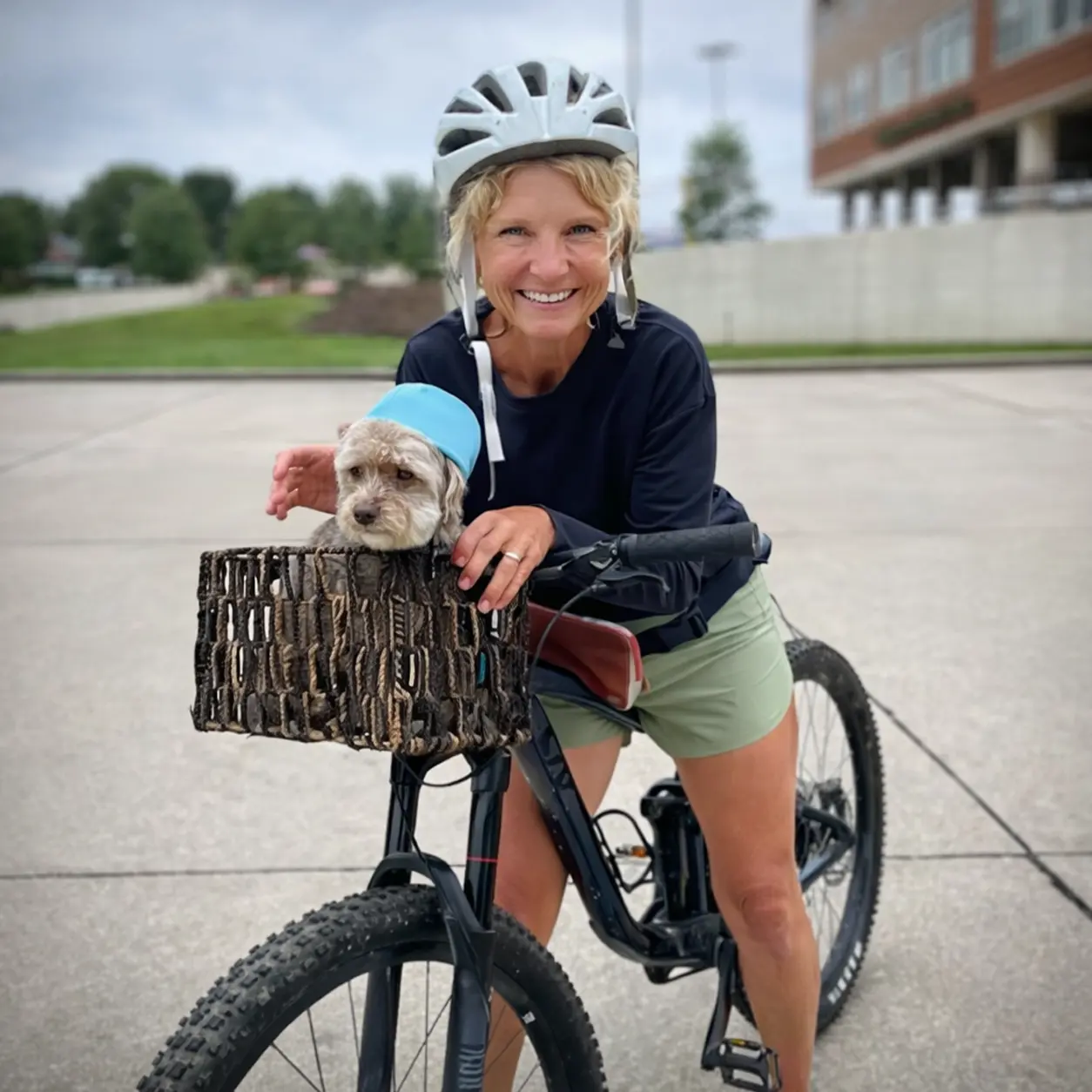 Shelly G riding a bicycle with a basket, smiling outdoors on a paved path.