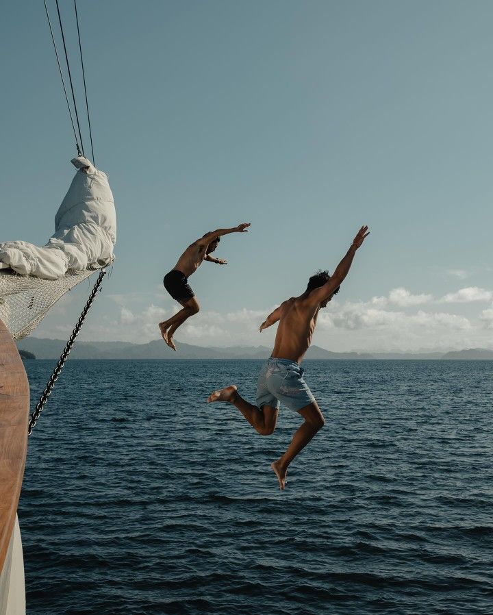 Two men jumping off a sailboat into open ocean water under a cloudy sky