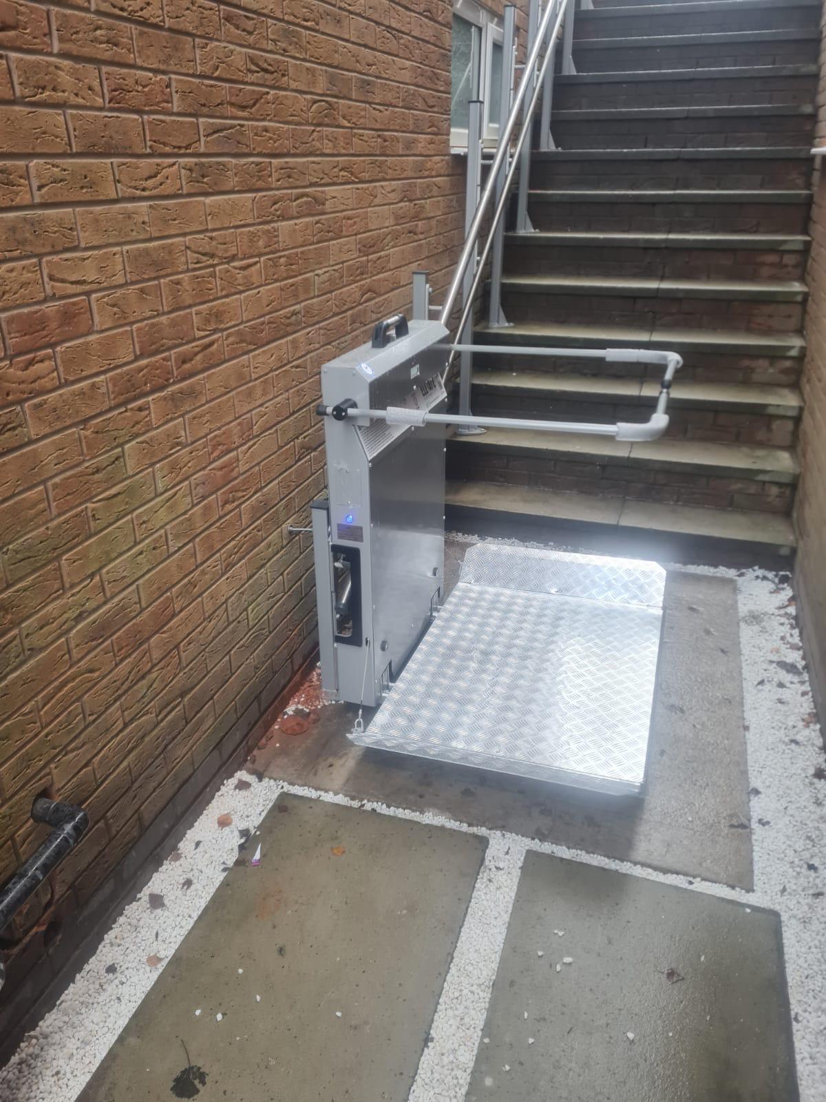 Inclined platform stairlift at lower landing position beside external steps — anti-slip chequerplate platform fully extended, wall-mounted drive unit with safety controls, outdoor UK installation