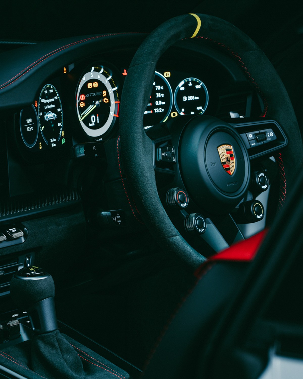Cockpit of a Porsche 911 GT3 RS (992) showing the Alcantara steering wheel with yellow centre marker and rotary drive-mode dials, the digital instrument cluster with track-screen display, and the Alcantara shifter.
