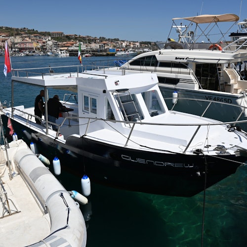 Un pequeño bote blanco llamado "GUENDREW" atracado en una marina, con personas a bordo y otras embarcaciones cercanas. En el fondo se ven los edificios del pueblo.