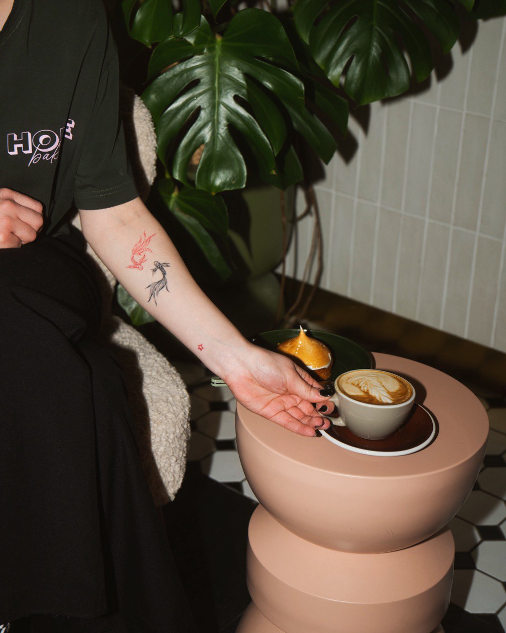 A person with colorful tattoos on their arm is seated next to a modern pink round table, reaching for a cup of frothy cappuccino, surrounded by lush green monstera leaves in a stylish setting.