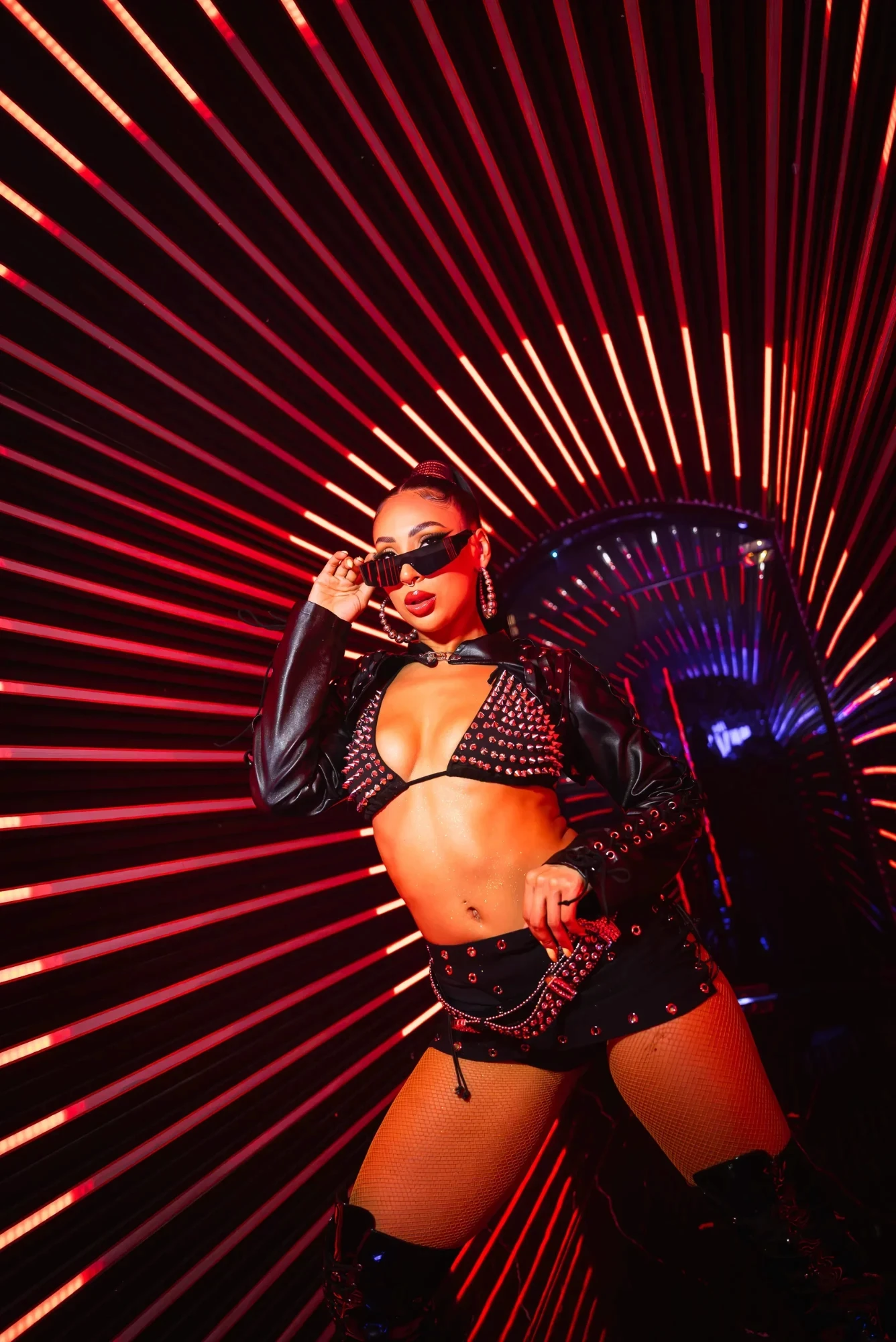 Performer in a studded black outfit and sunglasses posing against red light rays at The Myth Dubai