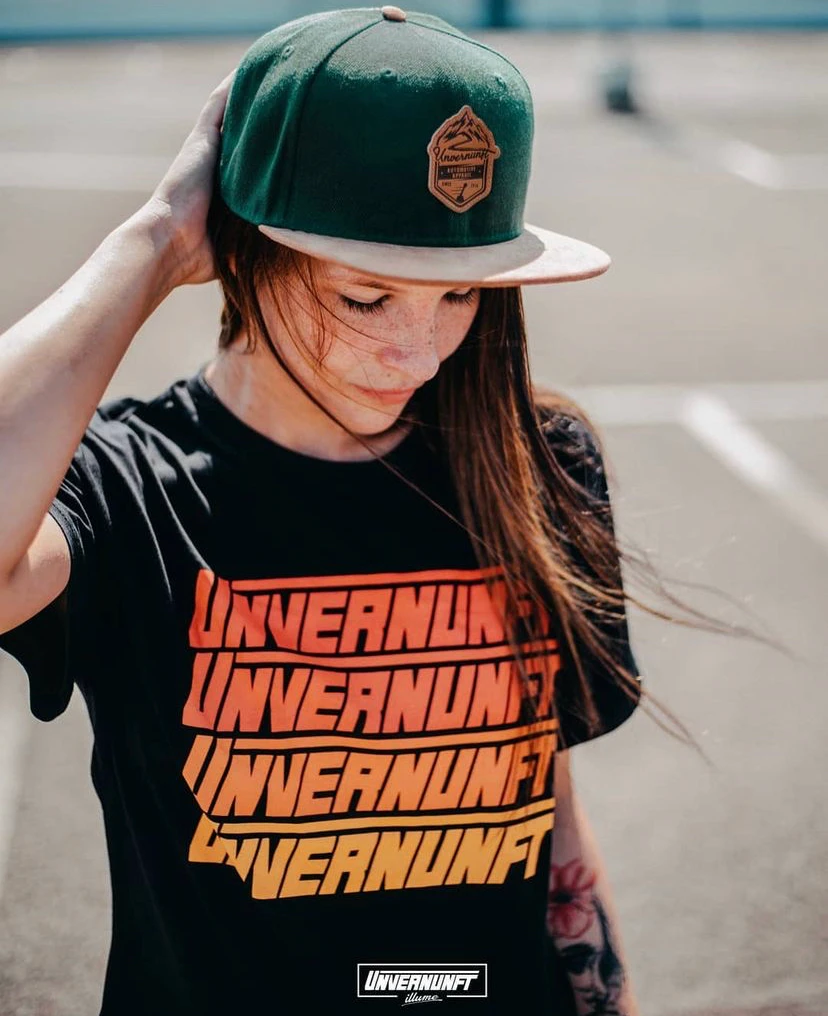 Model wearing Unvernunft black T-shirt with stacked orange gradient typography and branded green snapback with badge patch — apparel design by Slow Knife Design