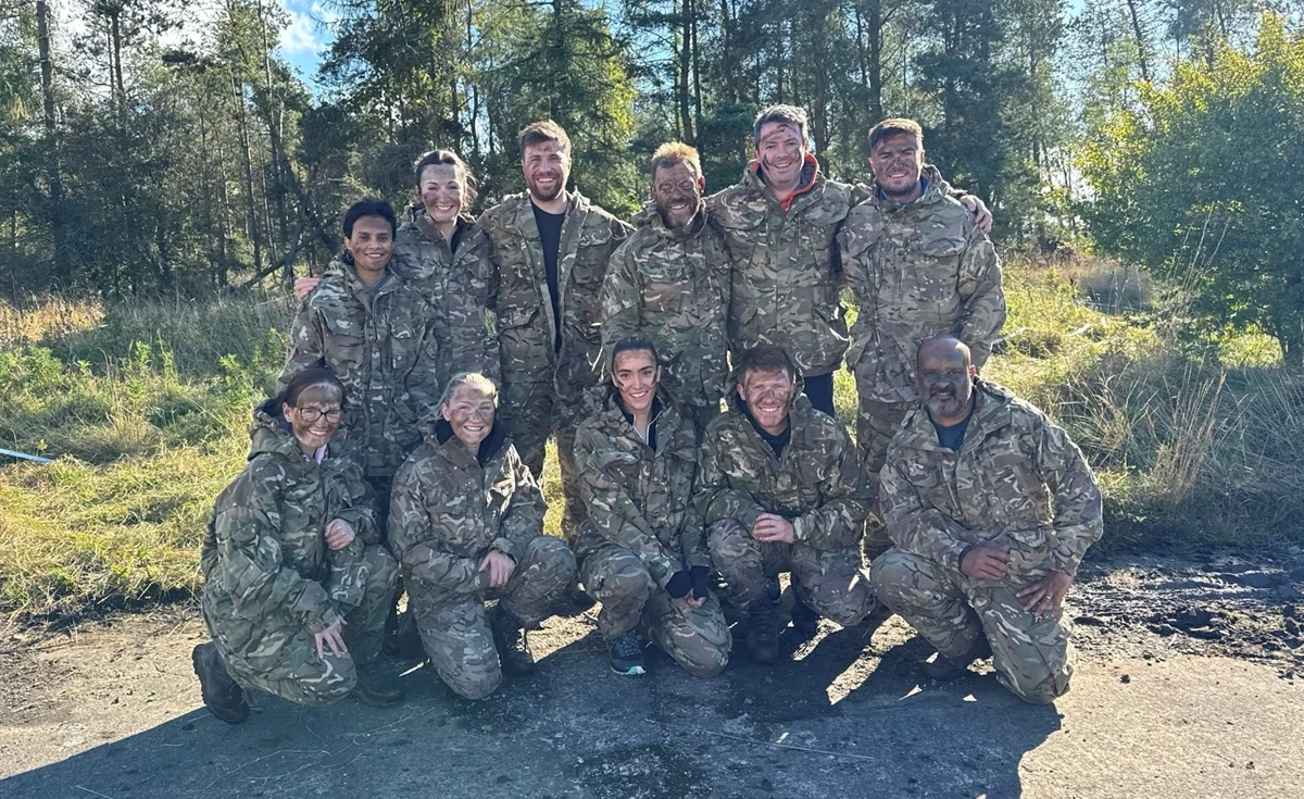 A group of people wearing military style camoflage jackets and trousers and face paint stand in a clearing in a forest smiling at the camera