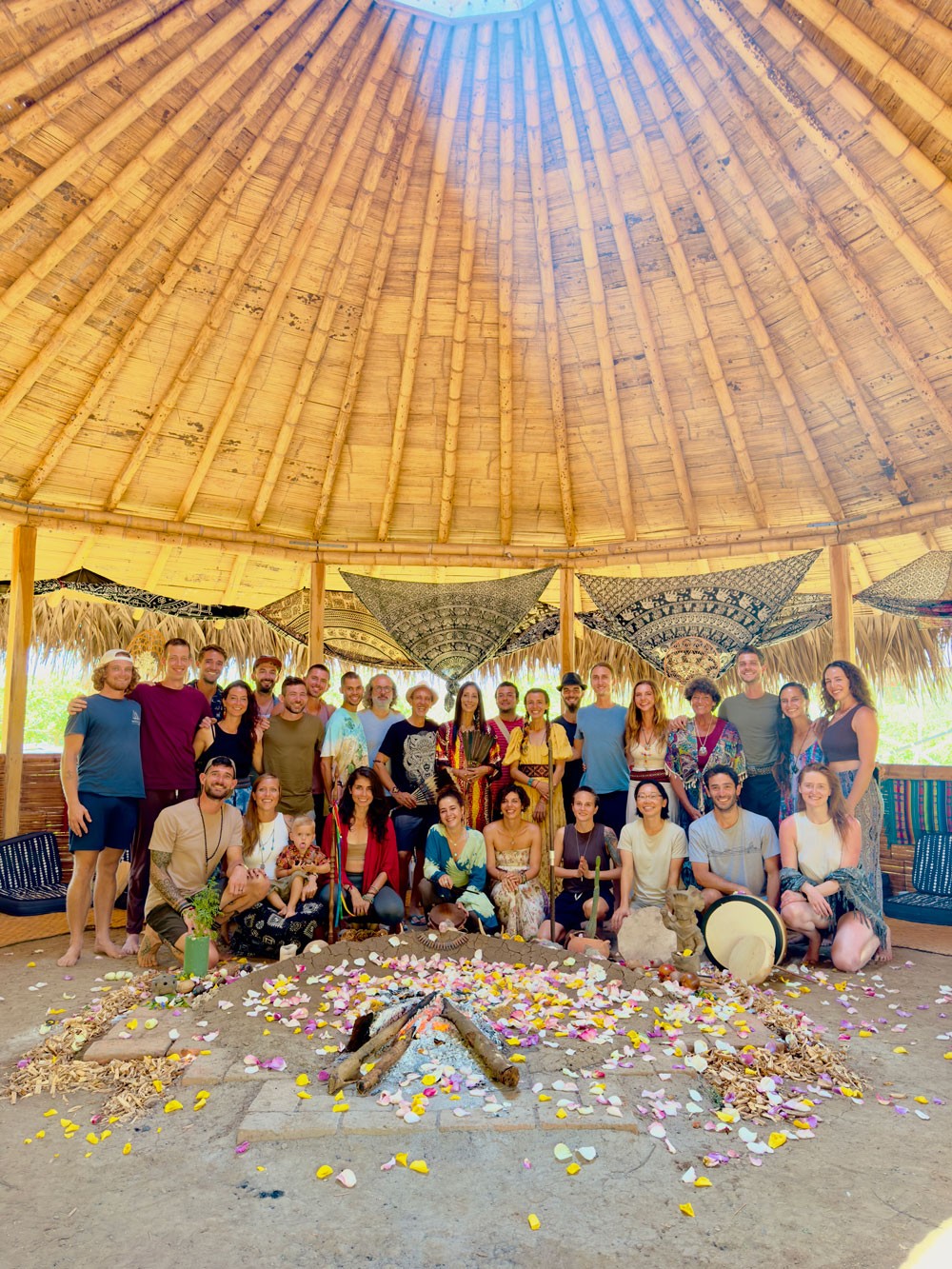 A group of yoga teacher students gather under a large, thatched dome, surrounding a circular arrangement of colorful flower petals and sticks. The scene conveys unity and joy.