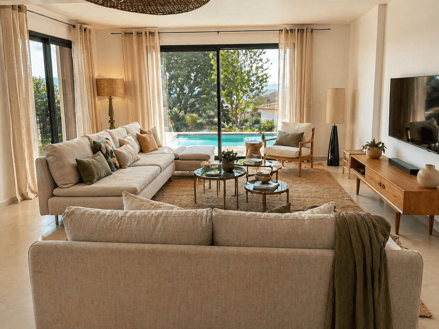 Salon d'une villa en Corse avec grand canapé d'angle beige, vue directe sur piscine, meuble TV en teck et suspensions en raphia