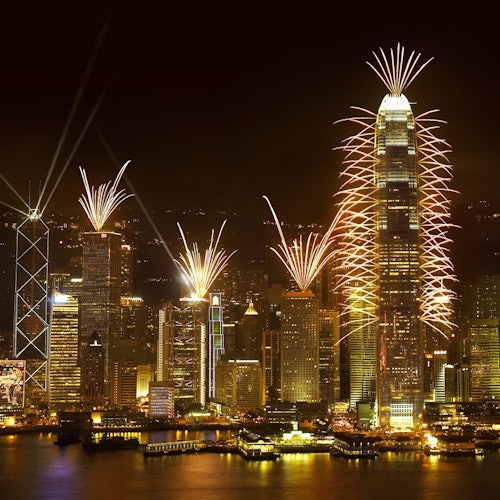 Éjszakai kilátás egy város látképére magas épületekkel, amelyeket tűzijáték díszít, és a látvány tükröződik az alattuk elterülő vízen.