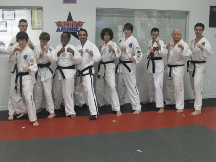 A group of ten karate students and instructors wearing white gis with black belts stand in a fighting stance lined up together on a red and gray mat in front of a dojo logo.