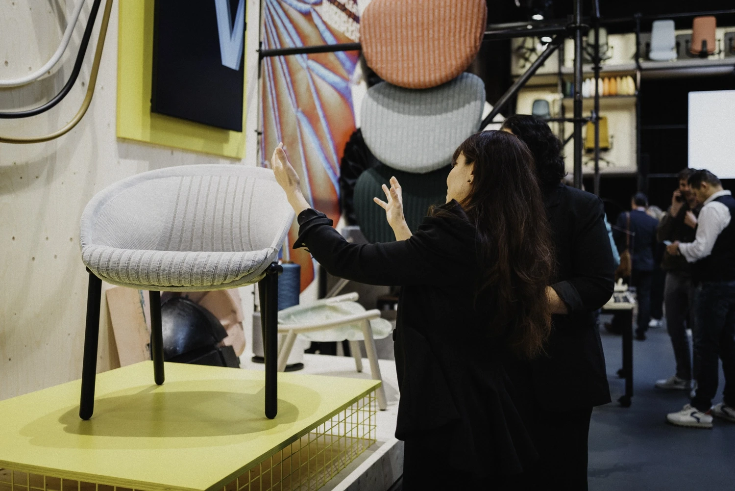 Visitor reaching toward a grey knit chair on a yellow display podium at the trade fair.