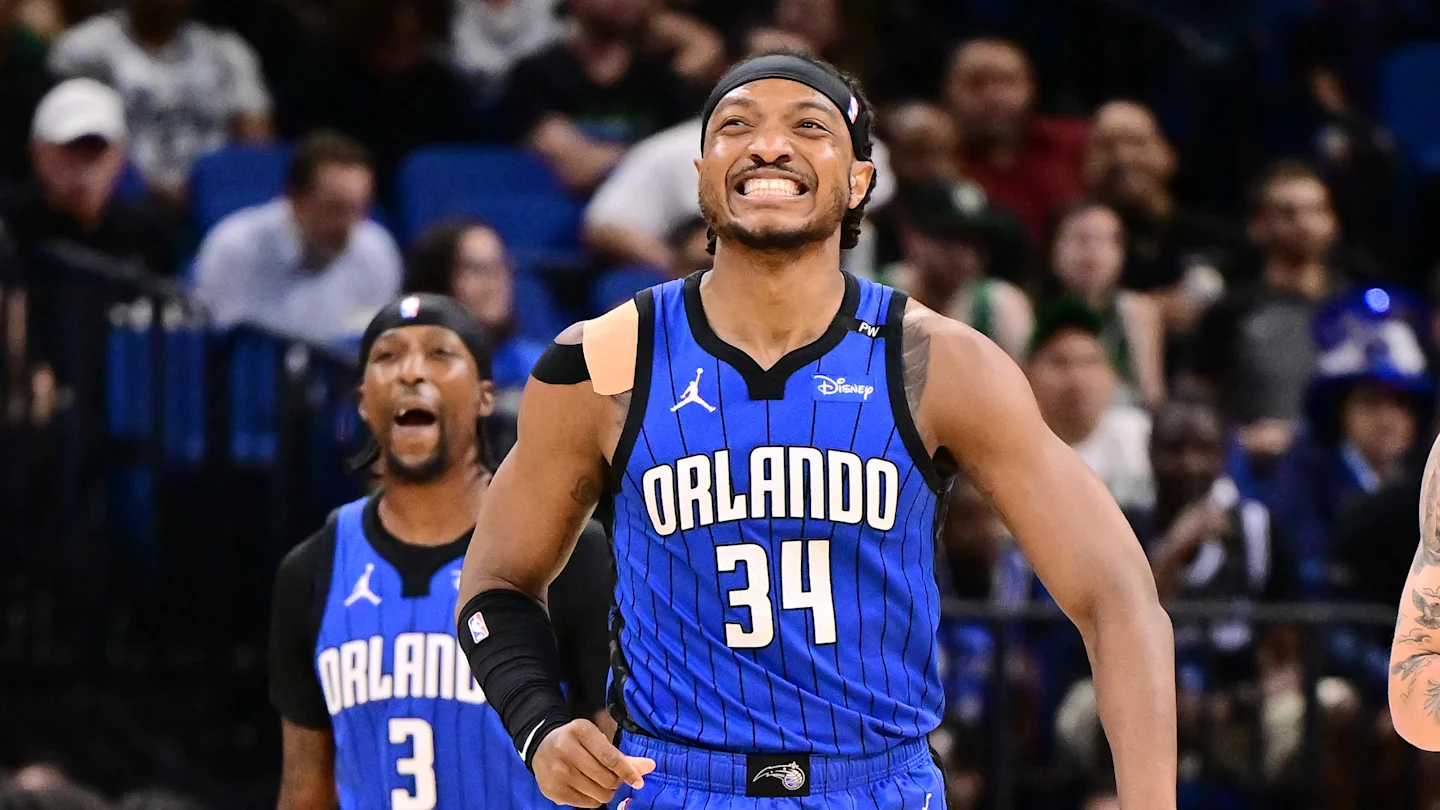 Orlando Magic player in a blue uniform smiling and celebrating during an NBA game with teammates in the background.