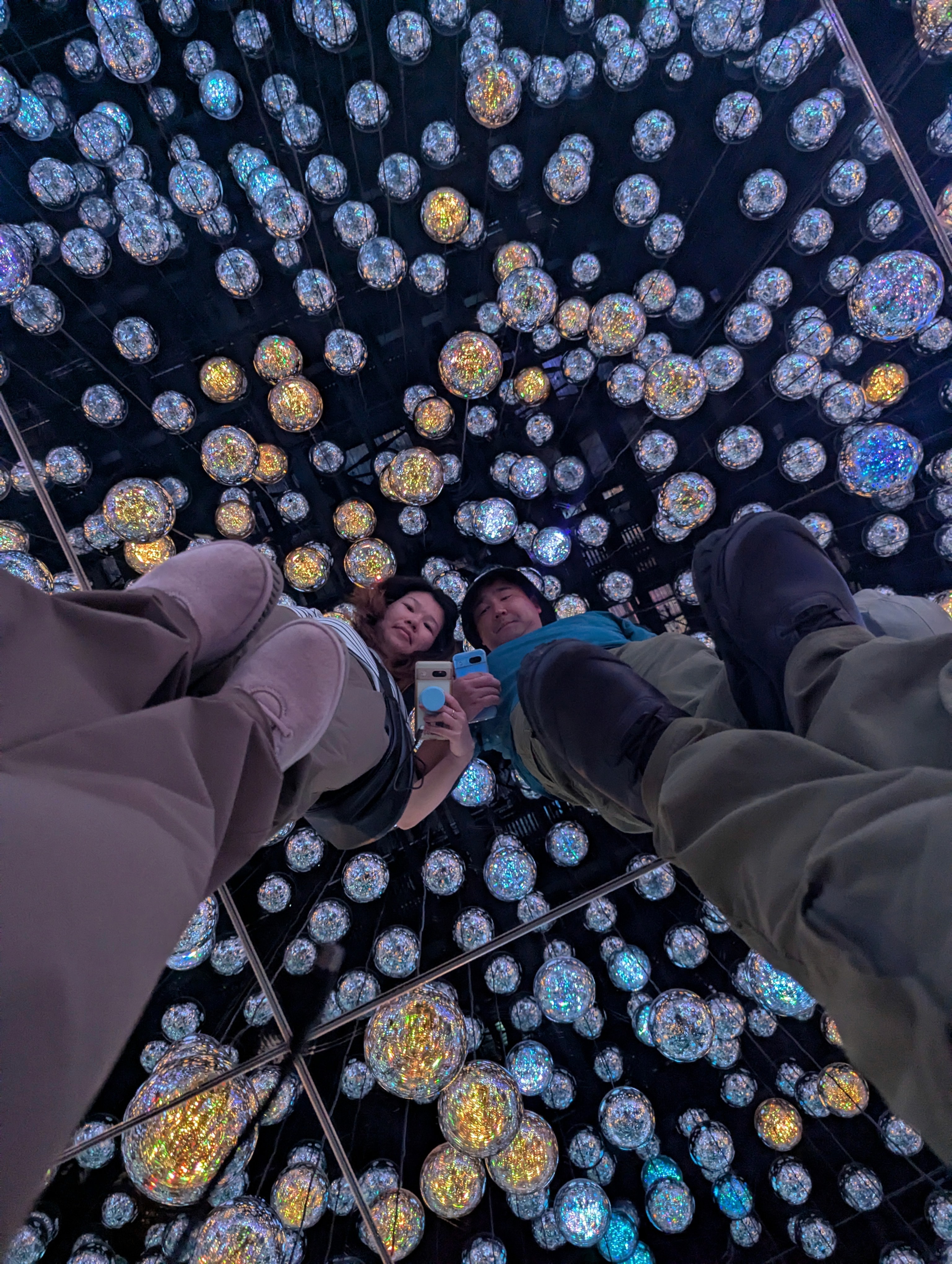 A groom in a dark suit carries a bride in a white dress across a sunny, grassy field. The couple shares a joyful, intimate moment with trees in the background.