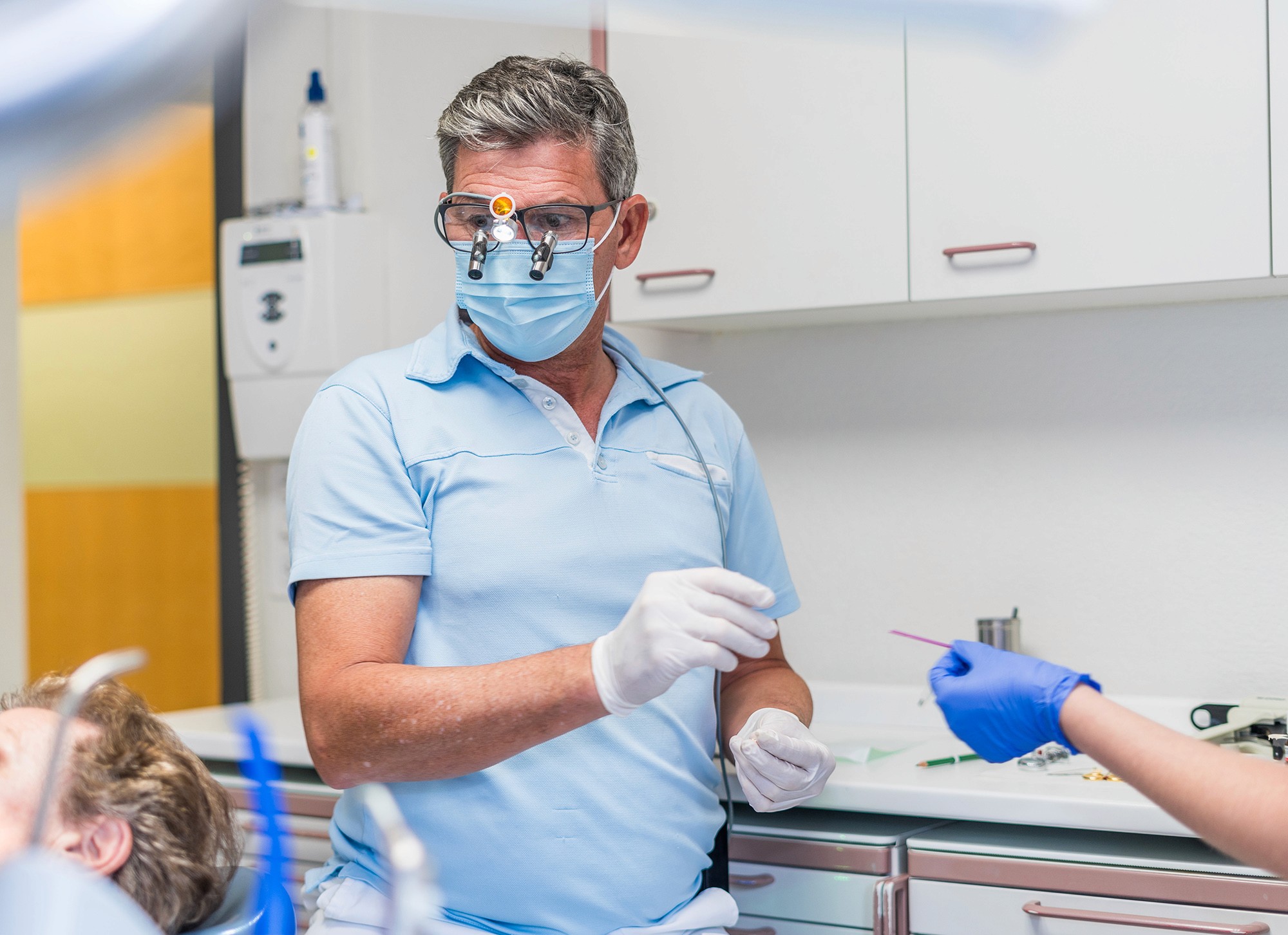 Zahnarzt Dr. Wimmer übergibt Instrument an Assistenz während laufender Behandlung am Patientenstuhl.