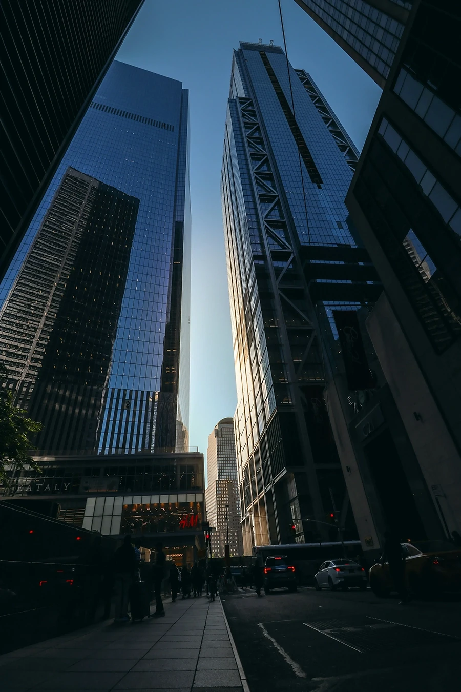 Street-level view looking up at tall modern skyscrapers with glass facades reflecting sunlight, cars, and pedestrians below