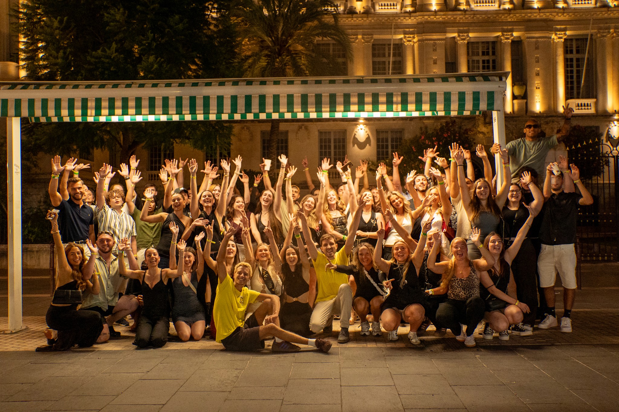 A group of international travelers laughing together at night in the streets of Nice, moving between bars with city lights around them.