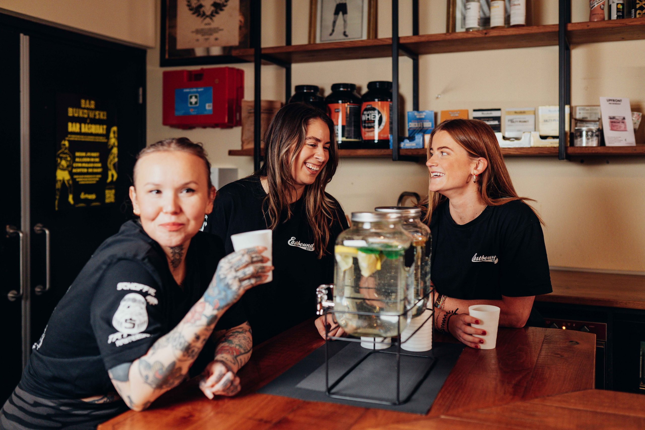 Three women are smiling and chatting behind a bar, surrounded by drinks and decor.