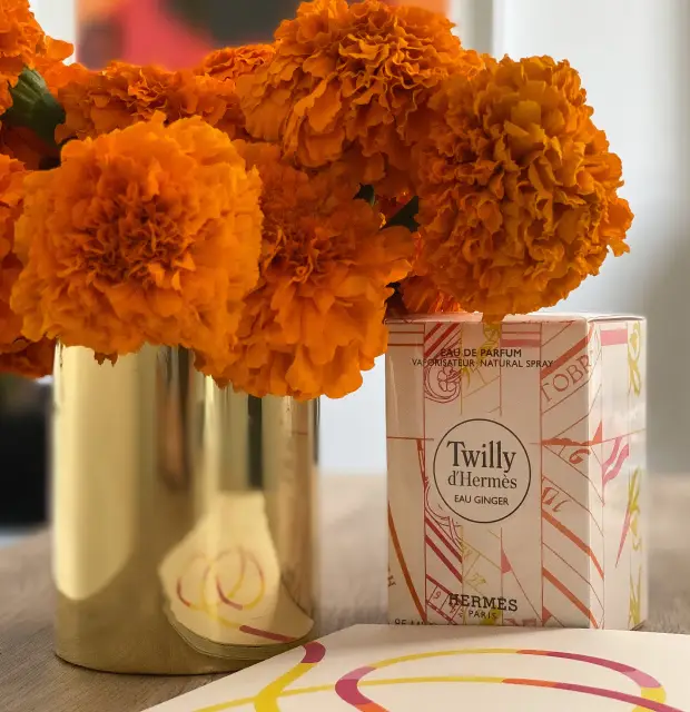 Ramo de flores de cempasúchil de color naranja intenso en un jarrón dorado junto a una caja del perfume Twilly d’Hermès Eau Ginger. La escena se completa con una hoja decorativa con líneas curvas en tonos rosa, amarillo y naranja, creando una composición cálida y elegante sobre una mesa de madera clara.