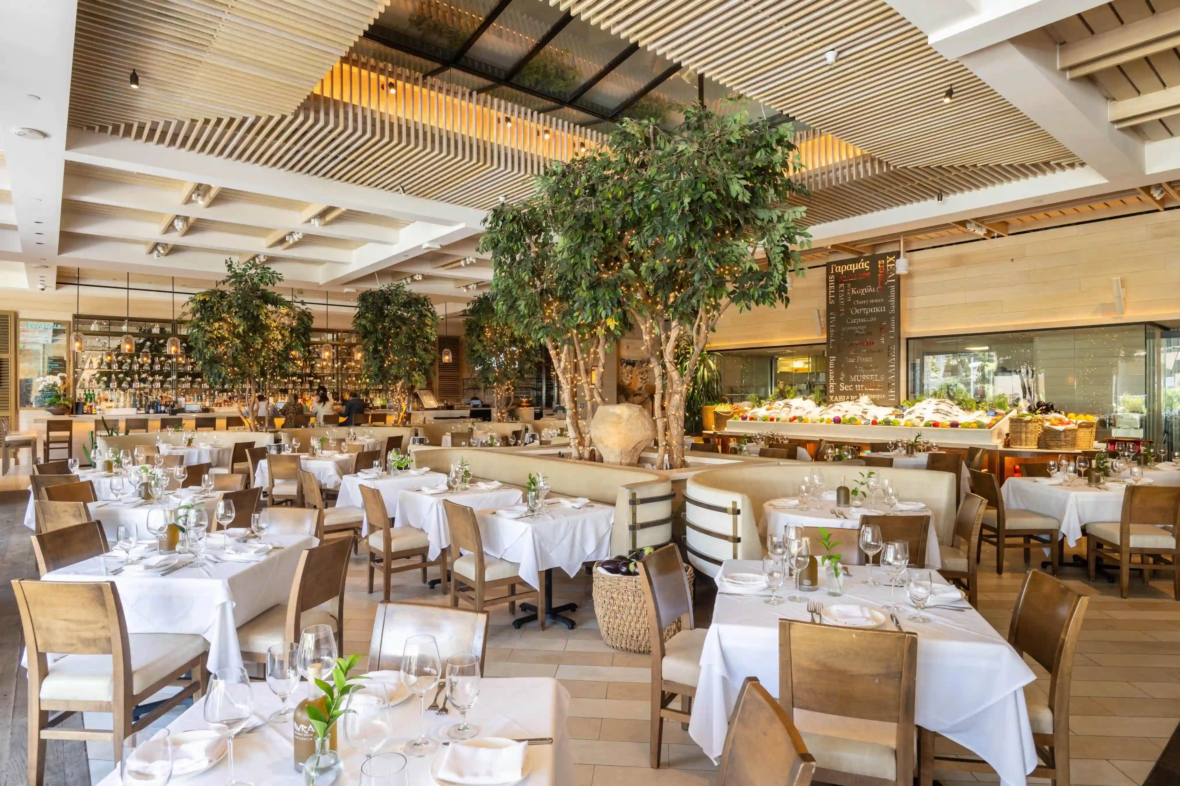 A spacious restaurant interior featuring white tablecloths, wooden furniture, and green plants.