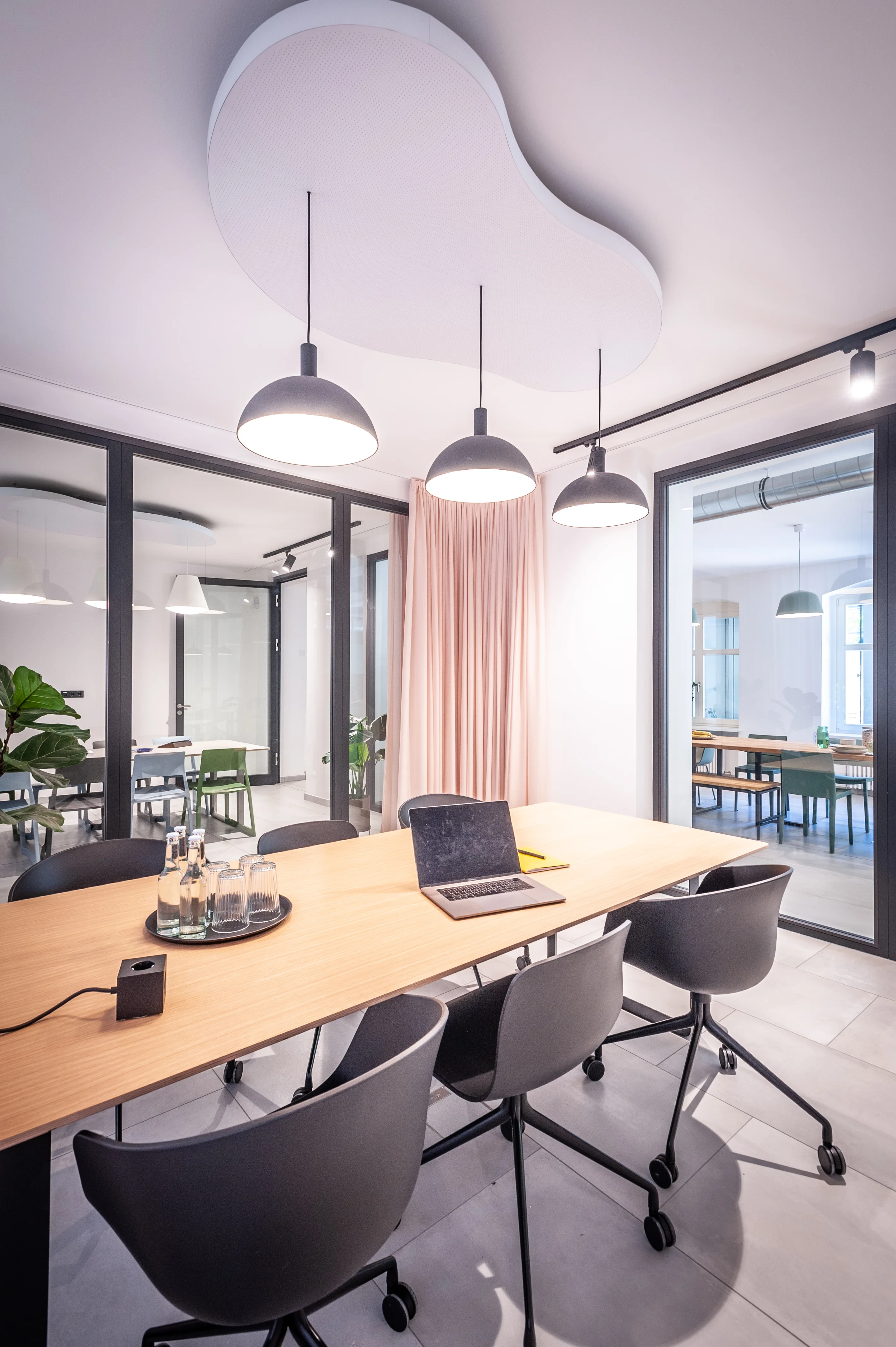 Conference room at EHEALTH-TEC with a cloud-shaped white acoustic ceiling panel, black dome pendant lights, an oak table with black shell chairs on castors and a blush curtain behind glass walls.