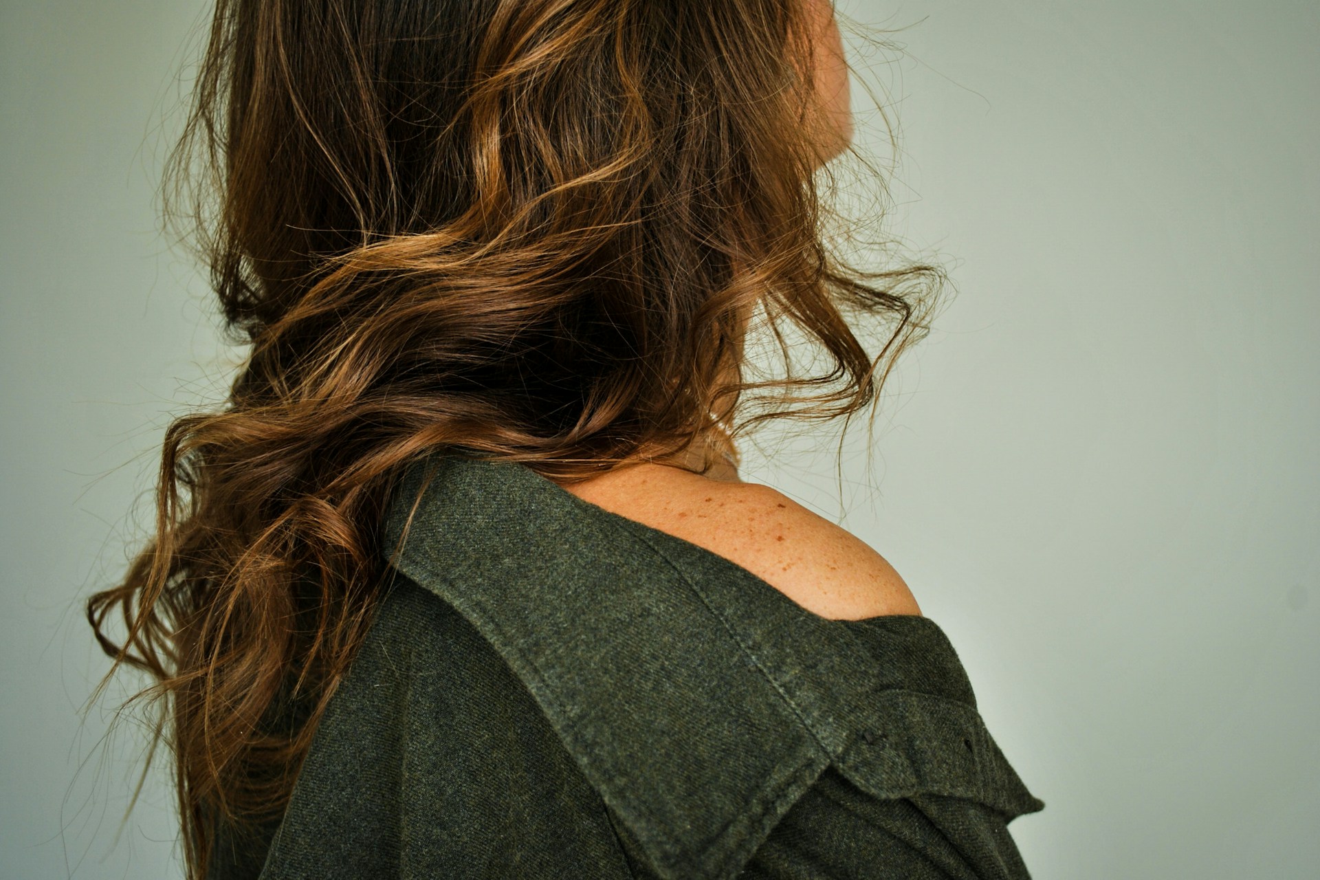 A side view showing the wavy brown hair and shoulder of a person wearing an olive green jacket.