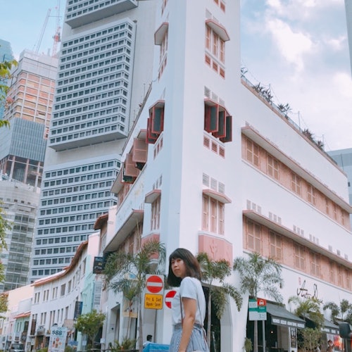 一位女士站在街角，背景是现代与旧式风格的高楼大厦，天空中多云少晴。