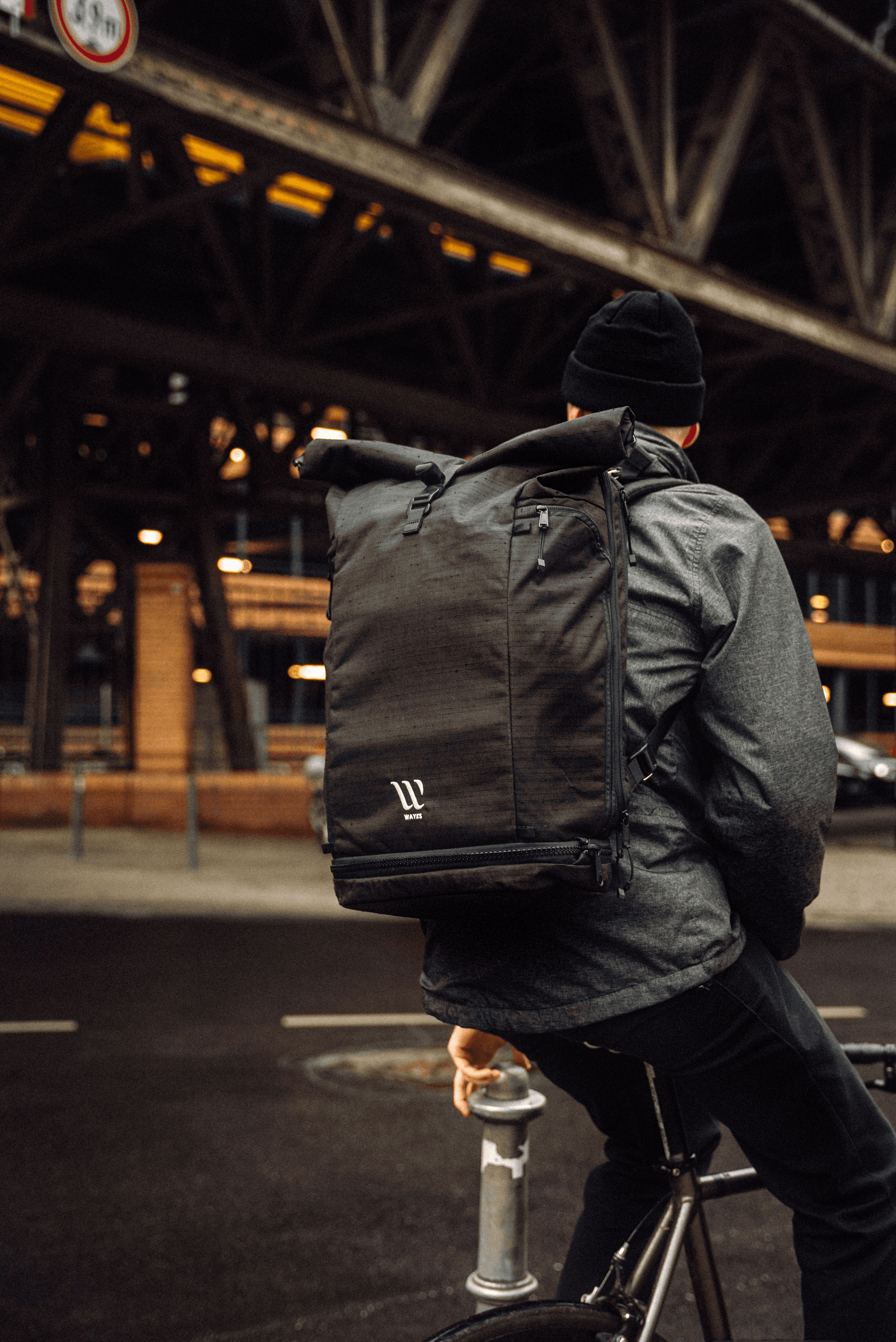 A person wearing dark clothing and carrying a large black backpack is sitting on a bicycle under an urban bridge, captured in a moody, industrial setting.