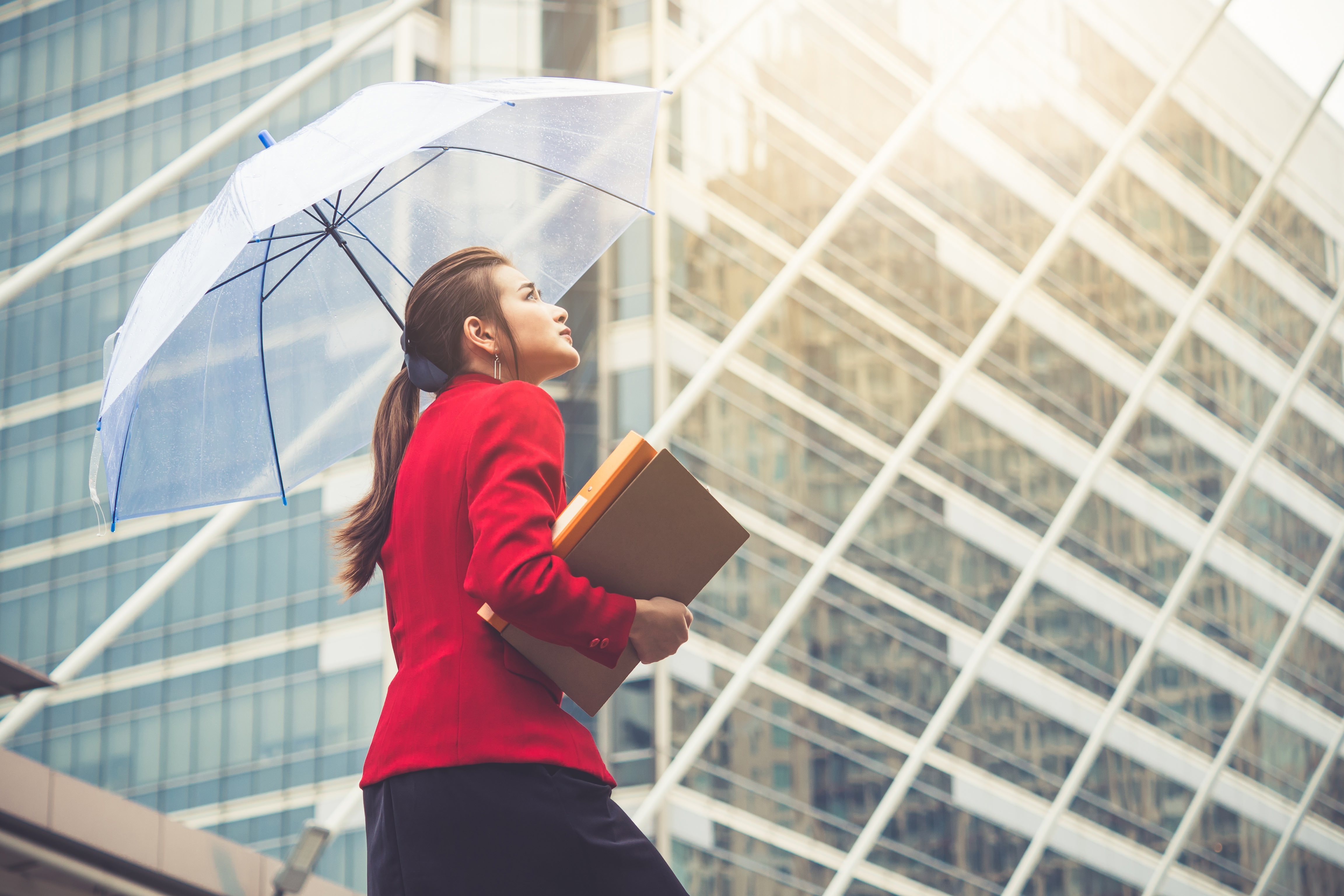 Businesswoman in a red blazer holding a transparent umbrella and folders, looking up at modern glass office buildings.