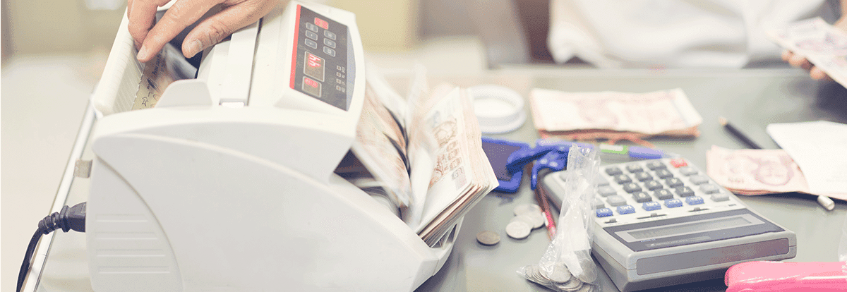 A close-up of a currency counting machine processing banknotes, accompanied by a calculator and coins, representing financial transactions or banking operations.