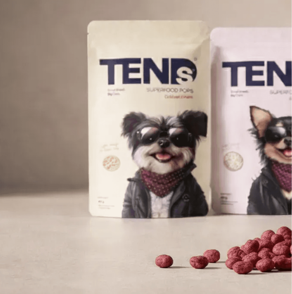 Two TENDS Superfood Pops pouches standing on a neutral surface with small round lotus seed snacks scattered in the foreground.