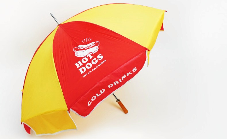 A red and yellow umbrella with a white hot dog logo and the words "HOT DOGS AND ICE COLD DRINKS" is shown against a white background. The words "COLD DRINKS" are printed along the bottom edge of the umbrella. The umbrella has a black handle and is angled slightly.