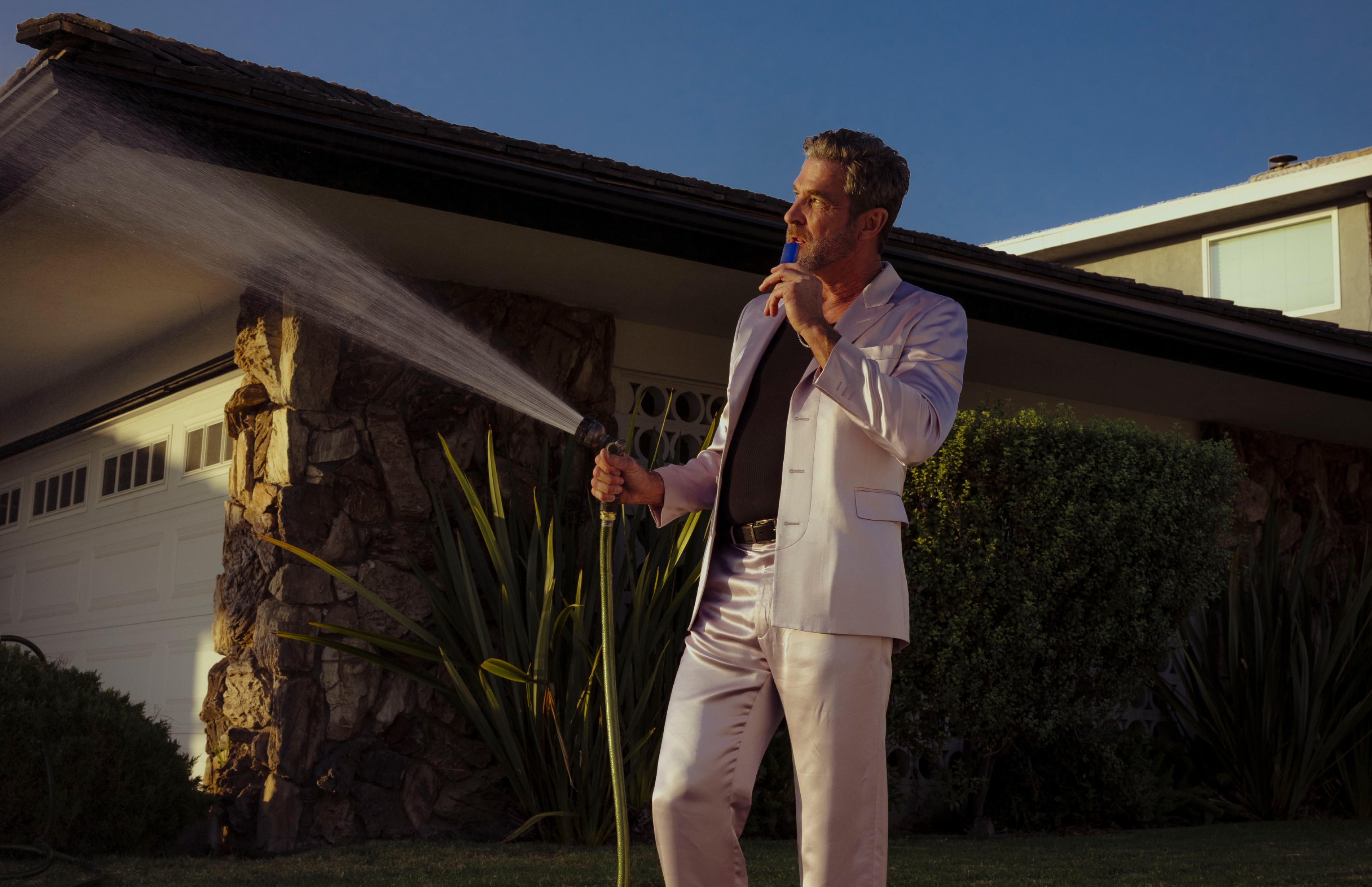 A man in a purple suit uses a PAX vaporizer as he waters the lawn.