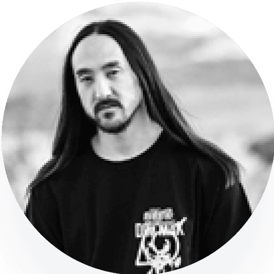 Steve Aoki, a man with long hair wearing a black shirt stands against a blurred background.