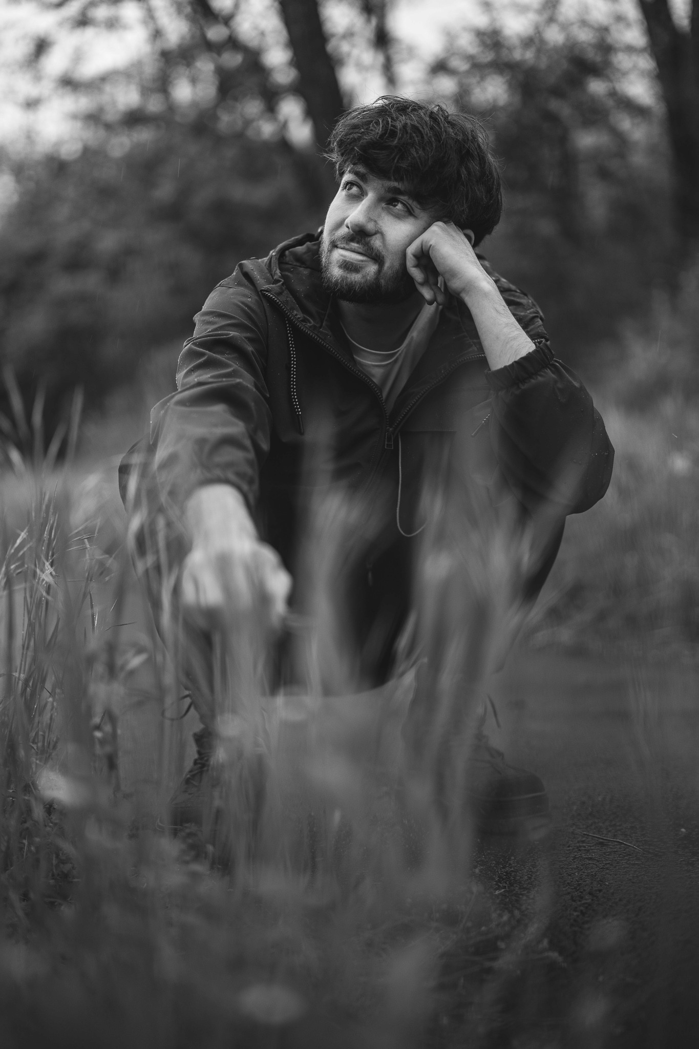Fiatal férfi ülve, gondolkodó pózban, esőkabátban, elmosódott fűszálakon keresztül, fekete-fehér természetfotón.