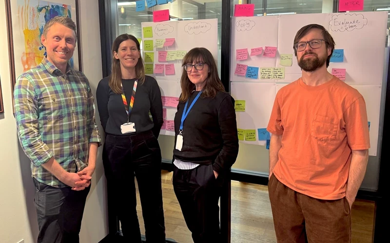 4 people stand in front of white boards covered in post it notes, a man in a checked shirt, a woman with long brown hair and dark clothes, a woman with dark brown long hair and a fringe in dark clothes and a blue lanyard, a many with short dark brown hair, a beard and an orange t shirt