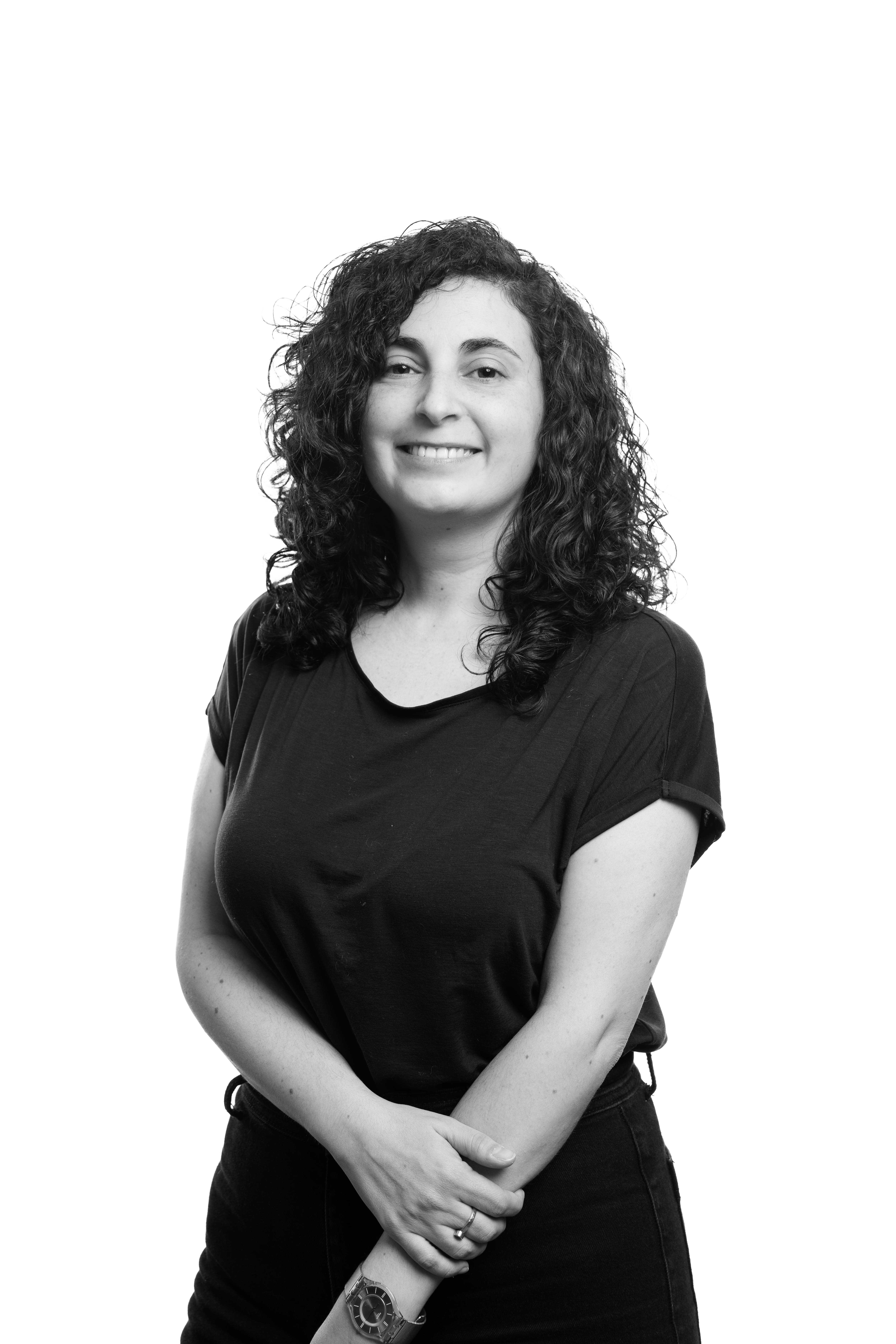 A woman with curly hair smiles warmly, wearing a dark top, against a plain white background. Her relaxed posture conveys a friendly, welcoming tone.
