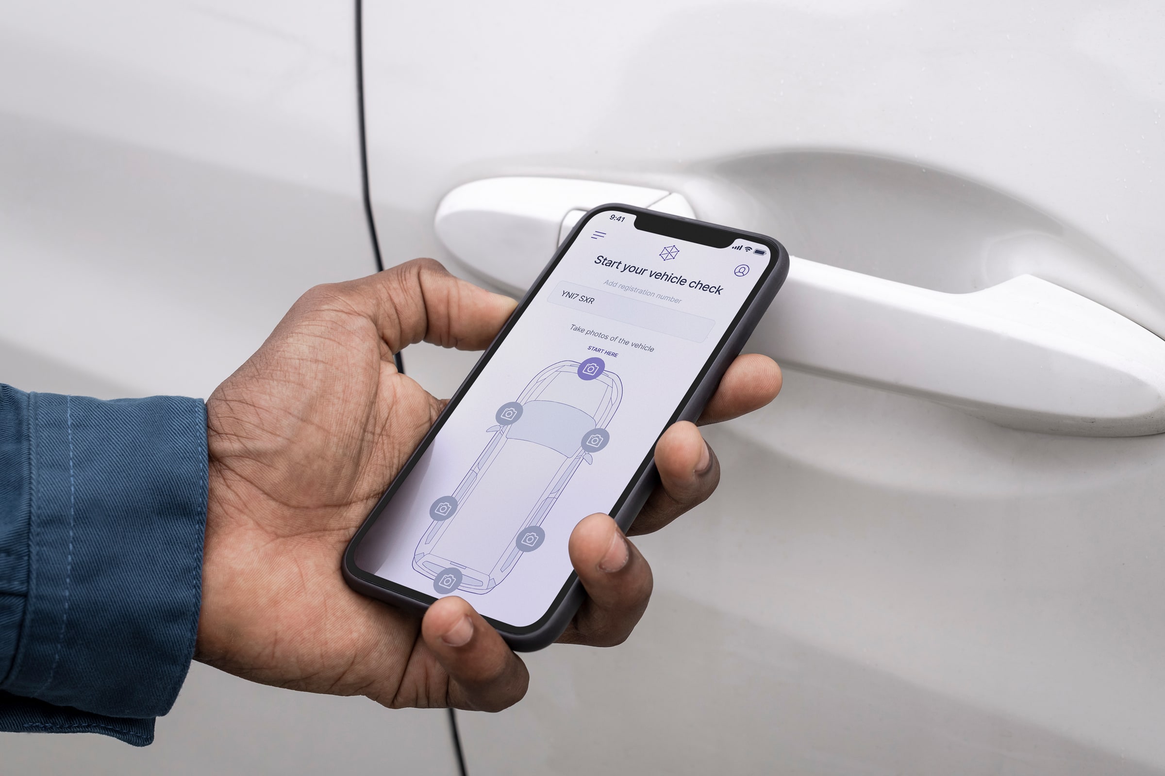 A hand holding an iPhone close to a white car door handle, with the DamageCheck app open showing the "Start your vehicle check" screen with a top-down vehicle diagram and camera icons marking six inspection positions.