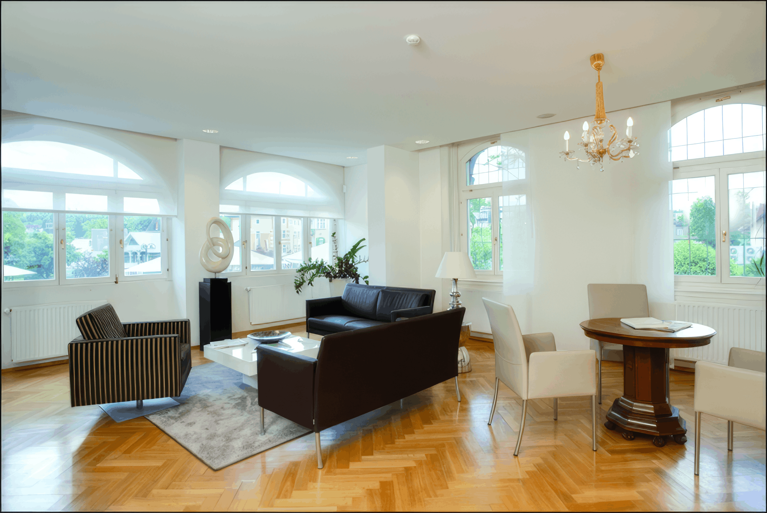 MW Interior - Heller repräsentativer Loungebereich in historischer Villa mit dunkelbraunem Ledersofa, Rundbogenfenstern, Fischgrätparkett und Kristalllüster" Beschreibung: Großzügiger Empfangs- und Besprechungsbereich mit dunklem Ledersofa, weißen Designstühlen, rundem Antiktisch und einer skulpturalen Kunstobjekt-Säule – Bogenfenster, Fischgrätparkett und ein goldener Lüster verleihen dem Raum eine unverwechselbare, elegante Atmosphäre.