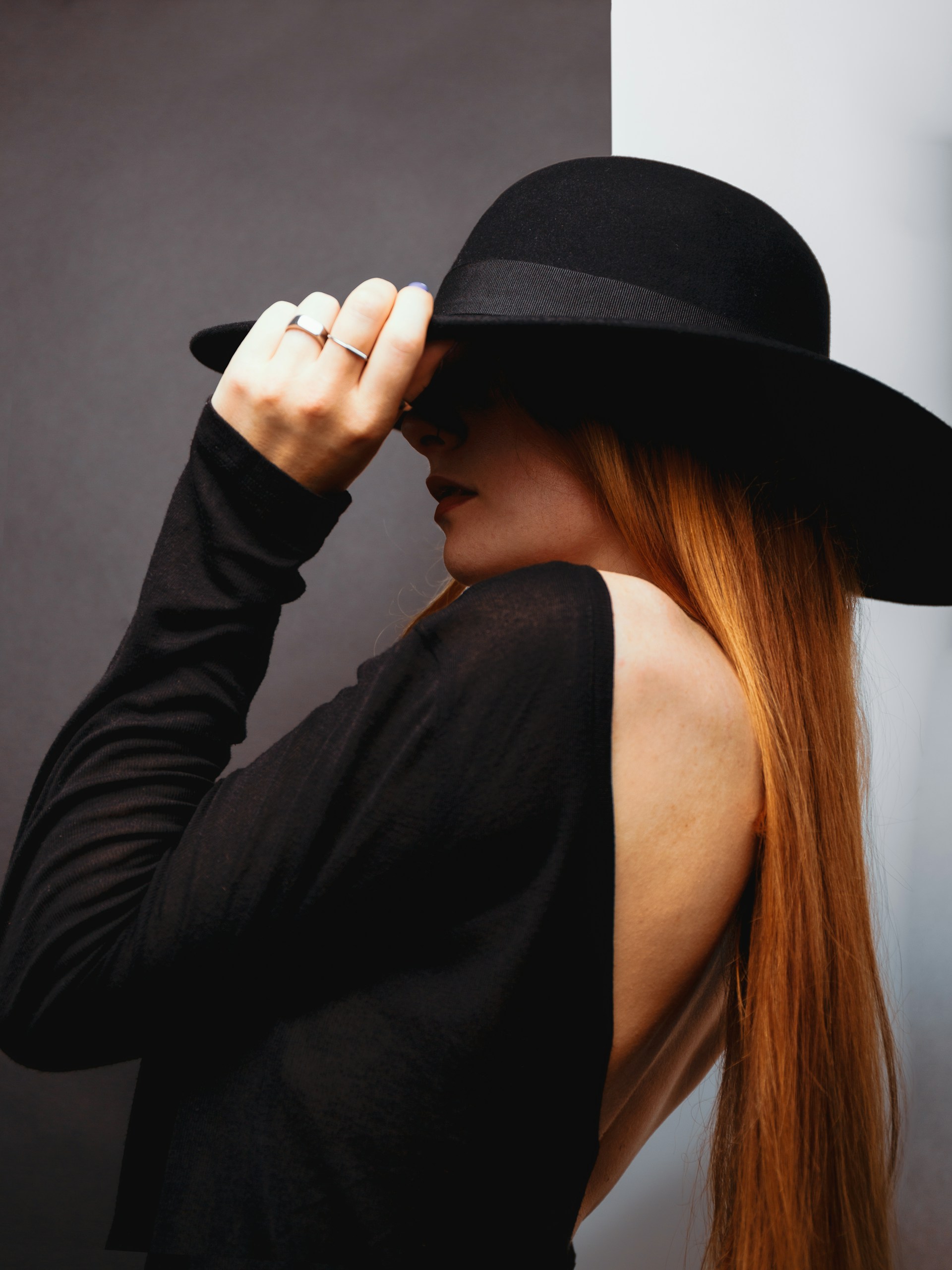 A woman with long red hair wearing a black wide-brimmed hat and a backless black top, partially obscured by shadows.