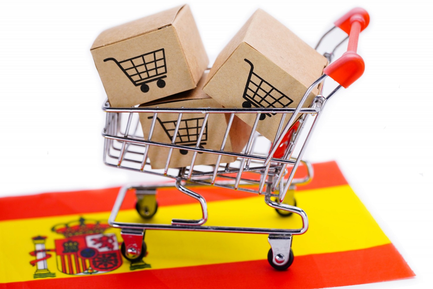 Un carrito de compras miniatura lleno de cajas de cartón con iconos de carritos de compra sobre un fondo que presenta la bandera de España.