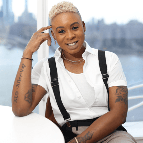 Ashleigh Warren, a smiling person with short hair and tattoos sitting at a table by a window, wearing a white shirt and black suspenders.
