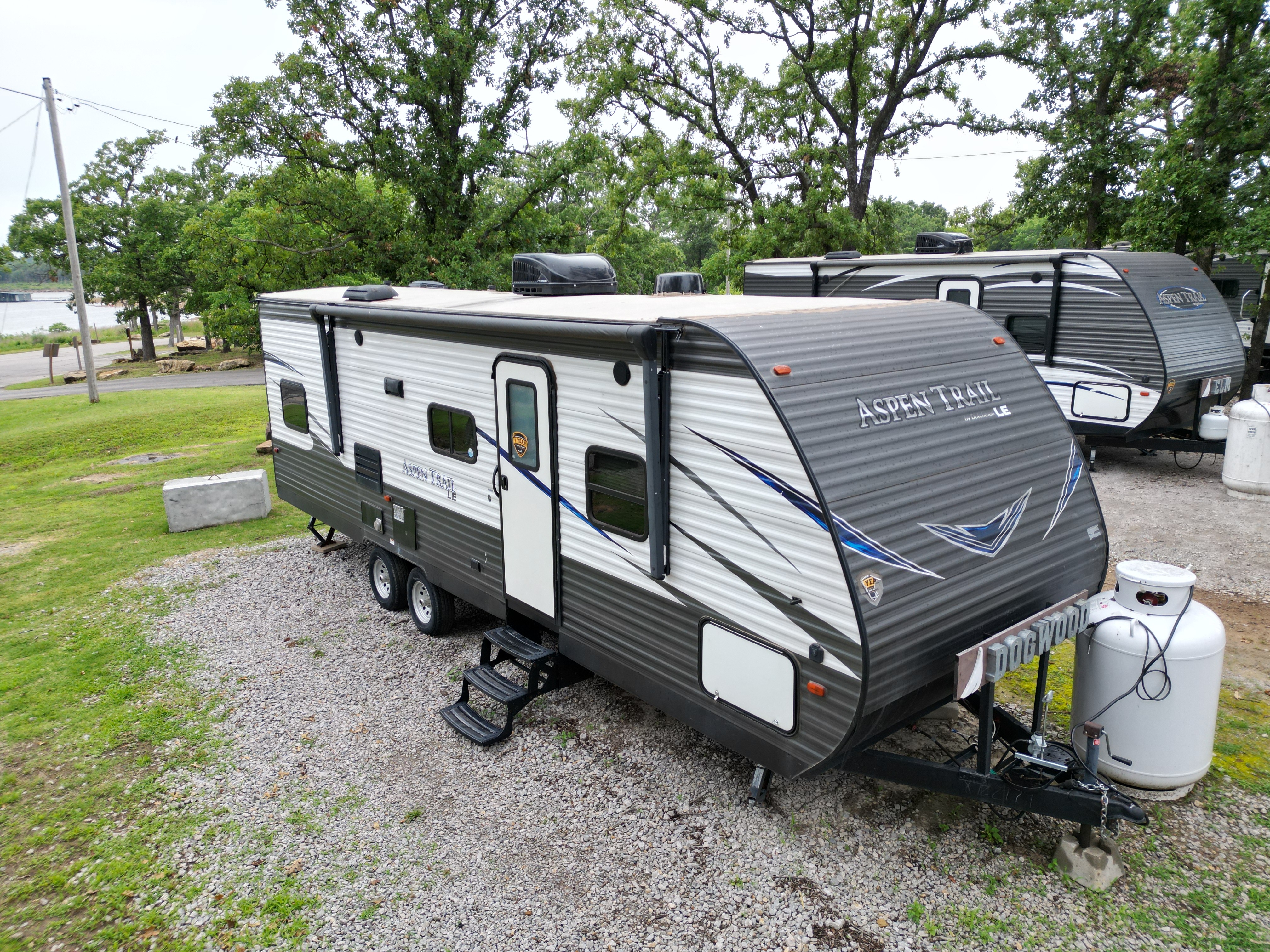 A well-maintained RV is parked on gravel with a backdrop of lush trees and a hint of water in the distance at what appears to be a campground or recreational area, in Keystone Harbor Marina.