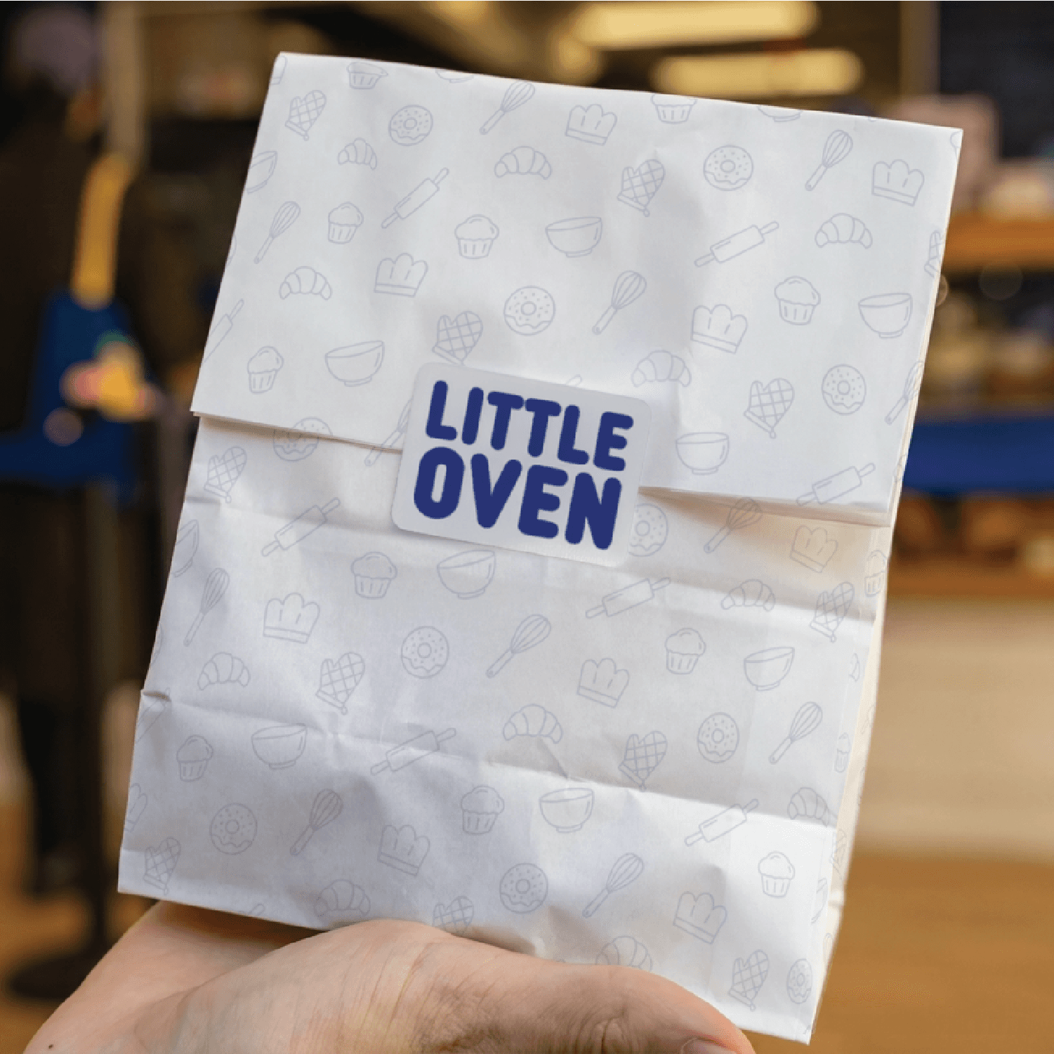 Hand holding a Little Oven pastry package printed with a subtle baking-icon pattern and indigo logo.