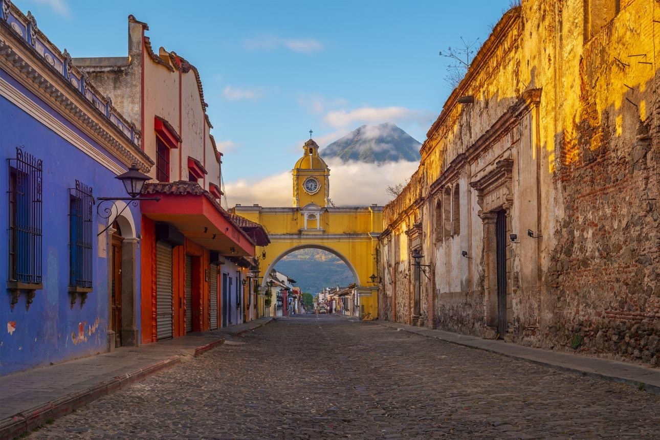 A picturesque cobblestone street in Antigua, Guatemala, features vibrant colonial buildings on either side and leads towards the iconic Santa Catalina Arch with a stunning volcano shrouded in clouds in the background.