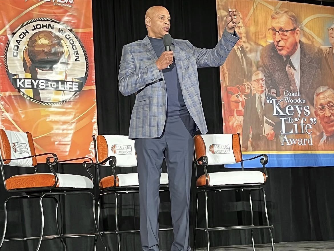 Clark Kellogg in a plaid blazer speaks on stage with a microphone. Behind him are empty chairs and banners featuring Coach John Wooden and the Keys to Life award.