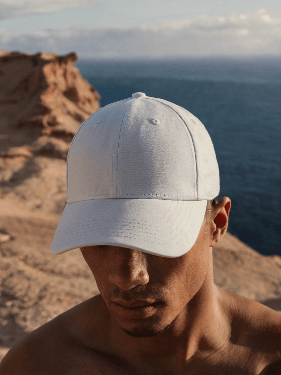 Man wearing a white baseball cap looking down, with a coastal cliff and the ocean in the background.