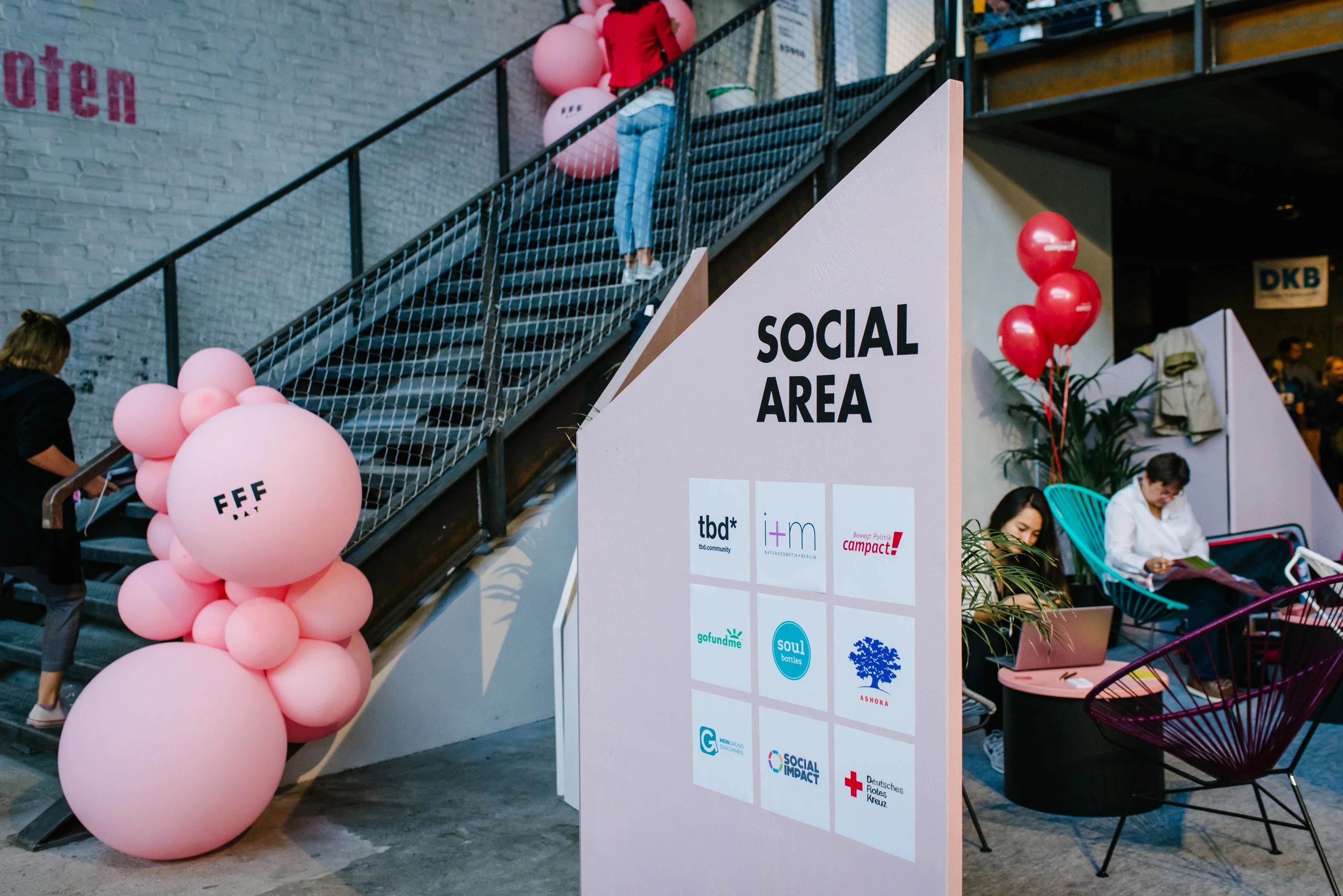 Social Area partner wall with sponsor logos at the FEMALE FUTURE FORCE DAY, pink balloon clusters on a steel staircase and women working in wire chairs.