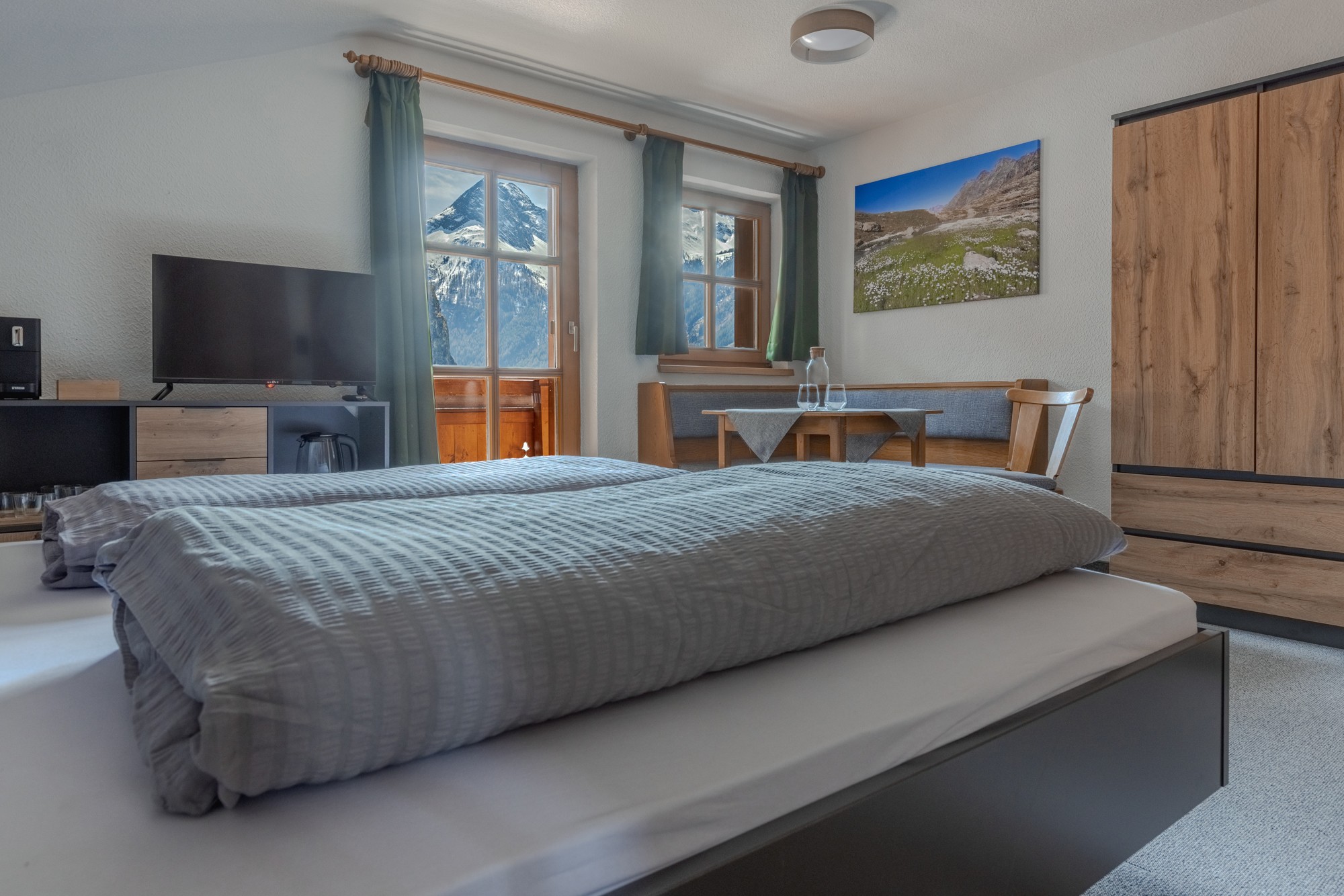 Ein gemütliches, modernes Schlafzimmer verfügt über ein Doppelbett mit grauer Bettwäsche, einen Holzschrank, einen kleinen Tisch mit Stühlen, einen an der Wand montierten Flachbildfernseher und ein großes Fenster mit atemberaubender Aussicht auf schneebedeckte Berge.
