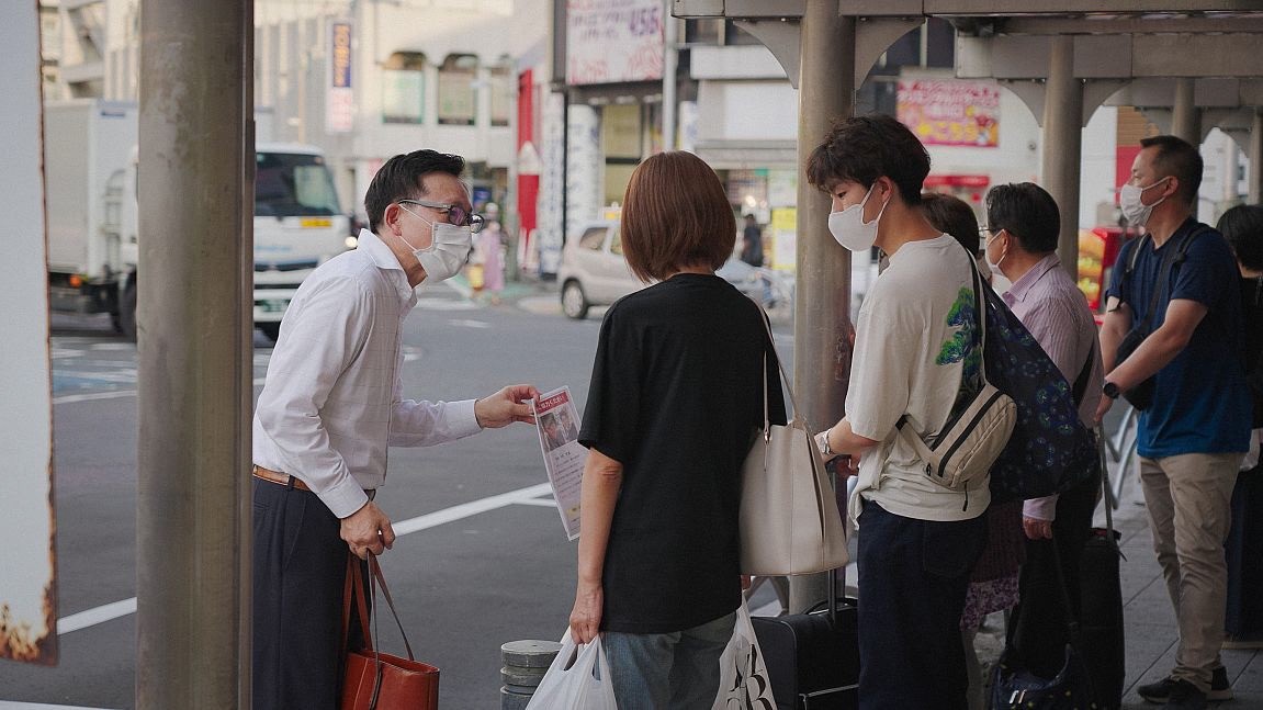 A group of people stands near a stall, engaging with a vendor in a bustling urban setting.