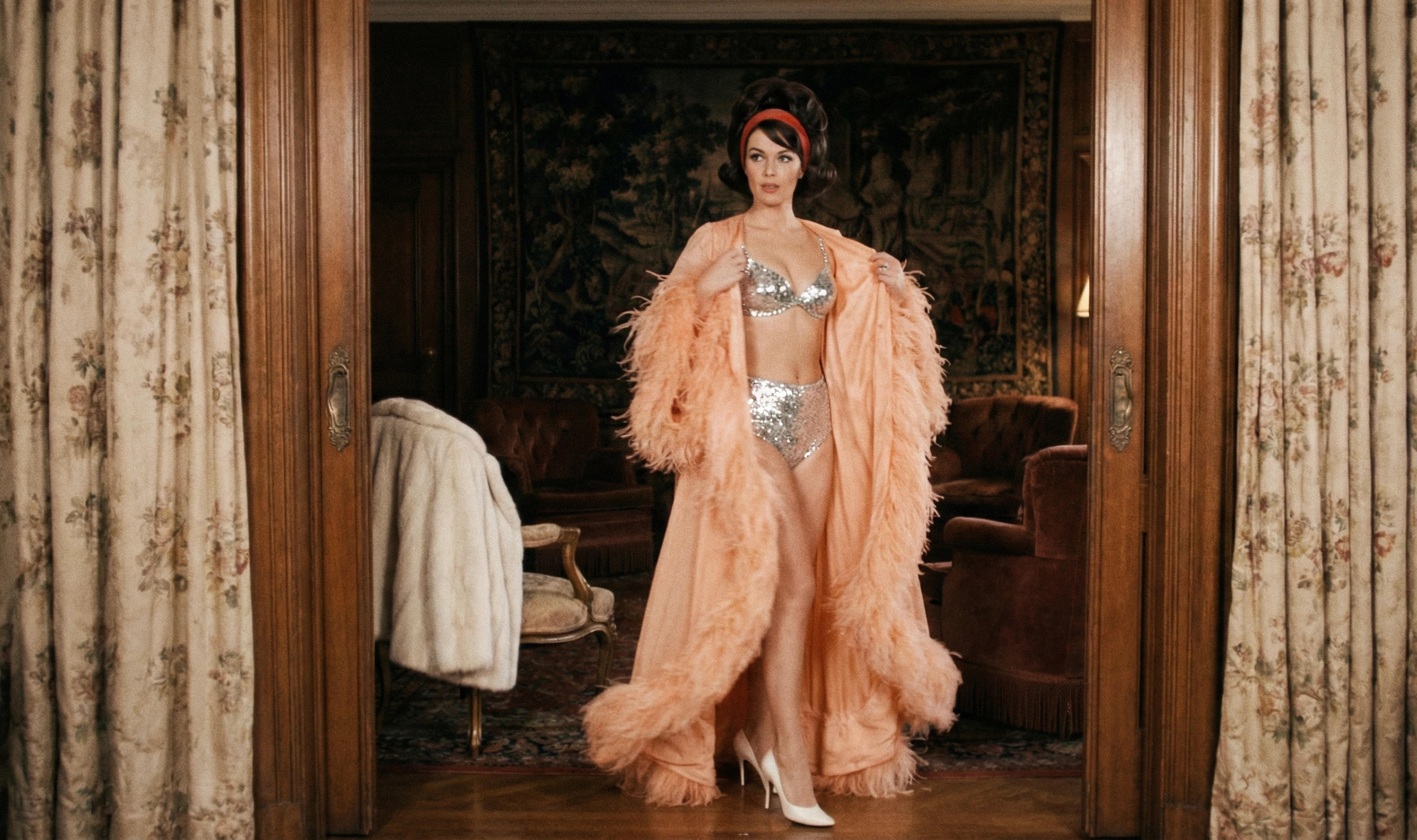 Woman in a silver embellished bra and briefs with a peach feathered robe in an ornate room.